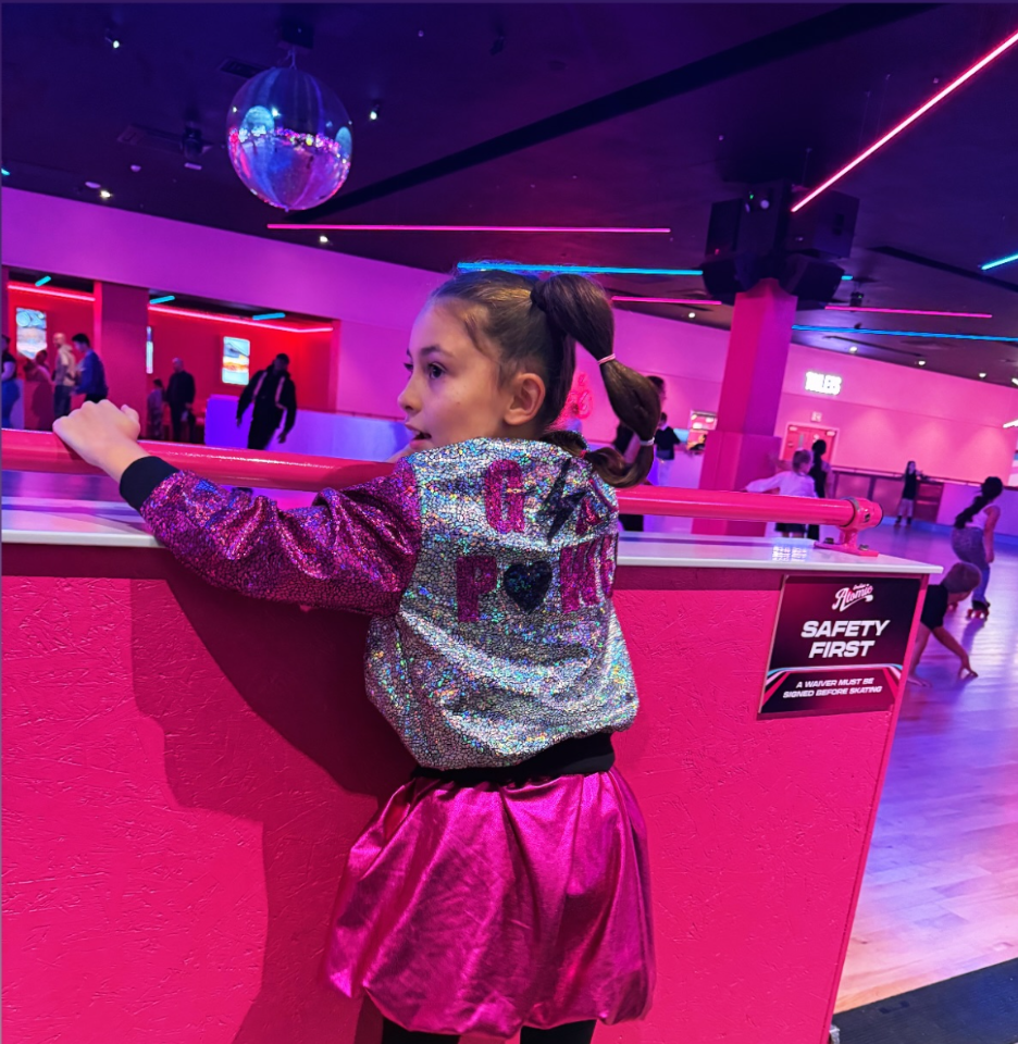 Young girl in a sparkly jacket and pink skirt standing in a vibrant, neon-lit indoor setting.