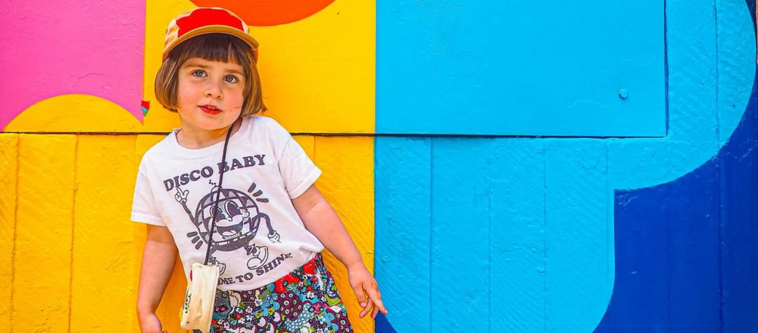 Child wearing a 'Disco Baby' t-shirt in front of a colorful wall with a large letter 'P'.