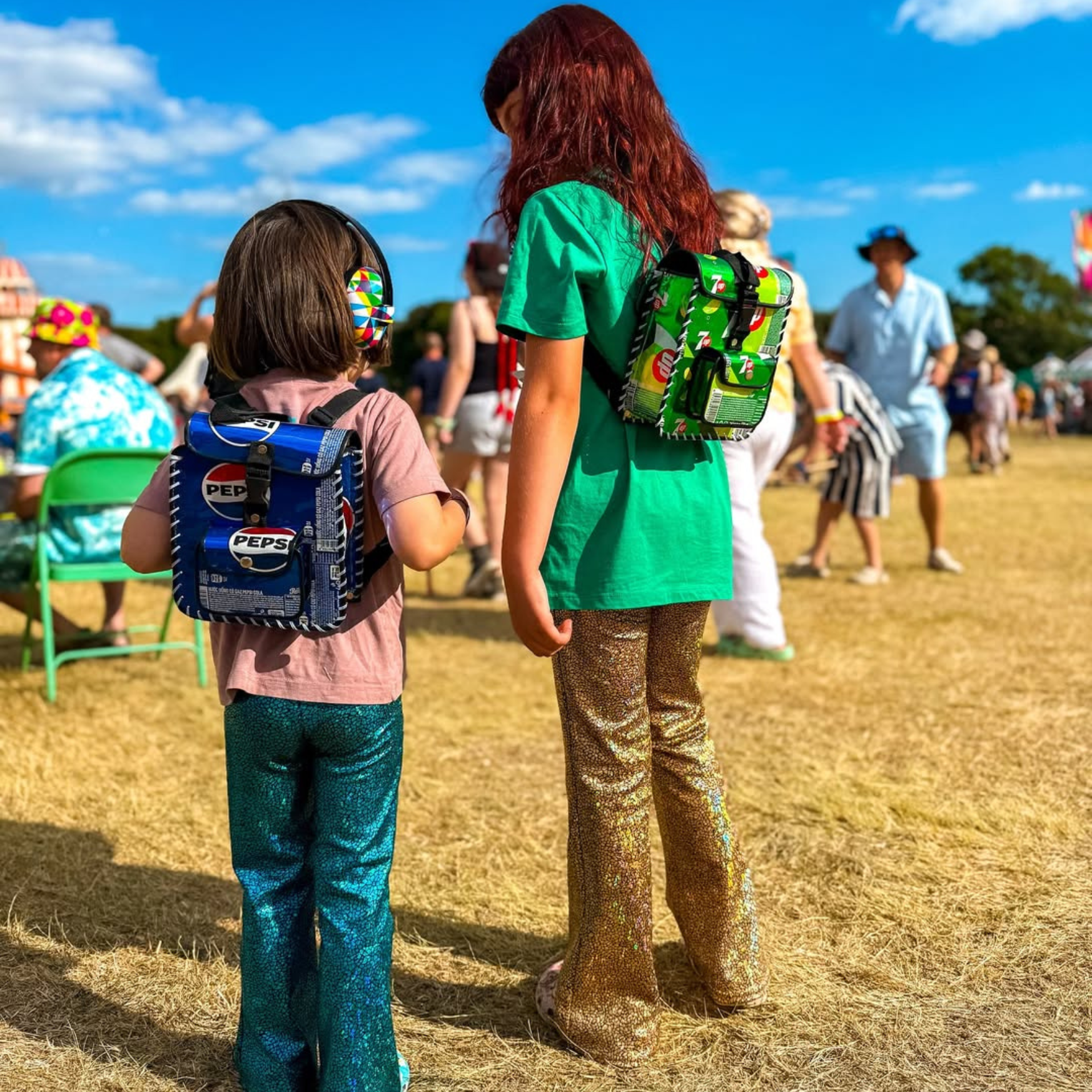 Two children with colorful backpacks standing on a grassy field with people in the background.