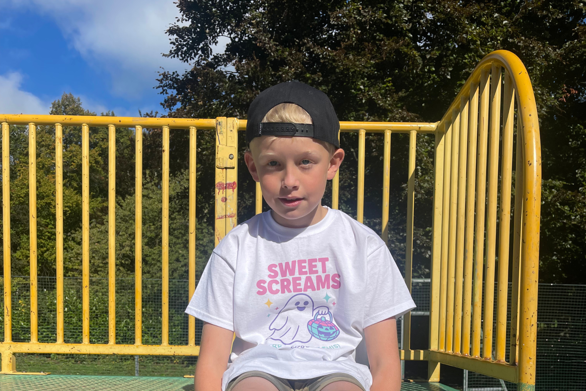 Child wearing a 'Sweet Screams' t-shirt sitting on a playground structure with trees in the background.