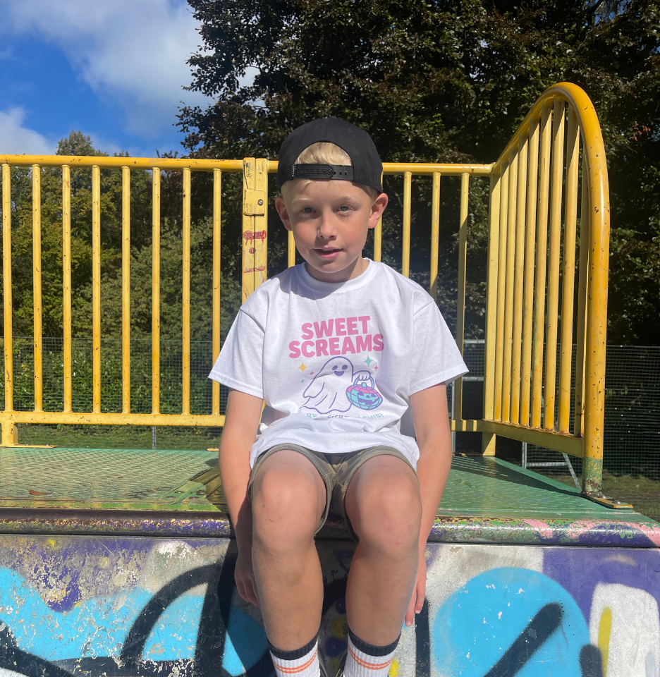 Child wearing a 'Sweet Screams' t-shirt sitting on a graffiti-covered wall with a yellow metal fence and trees in the background.