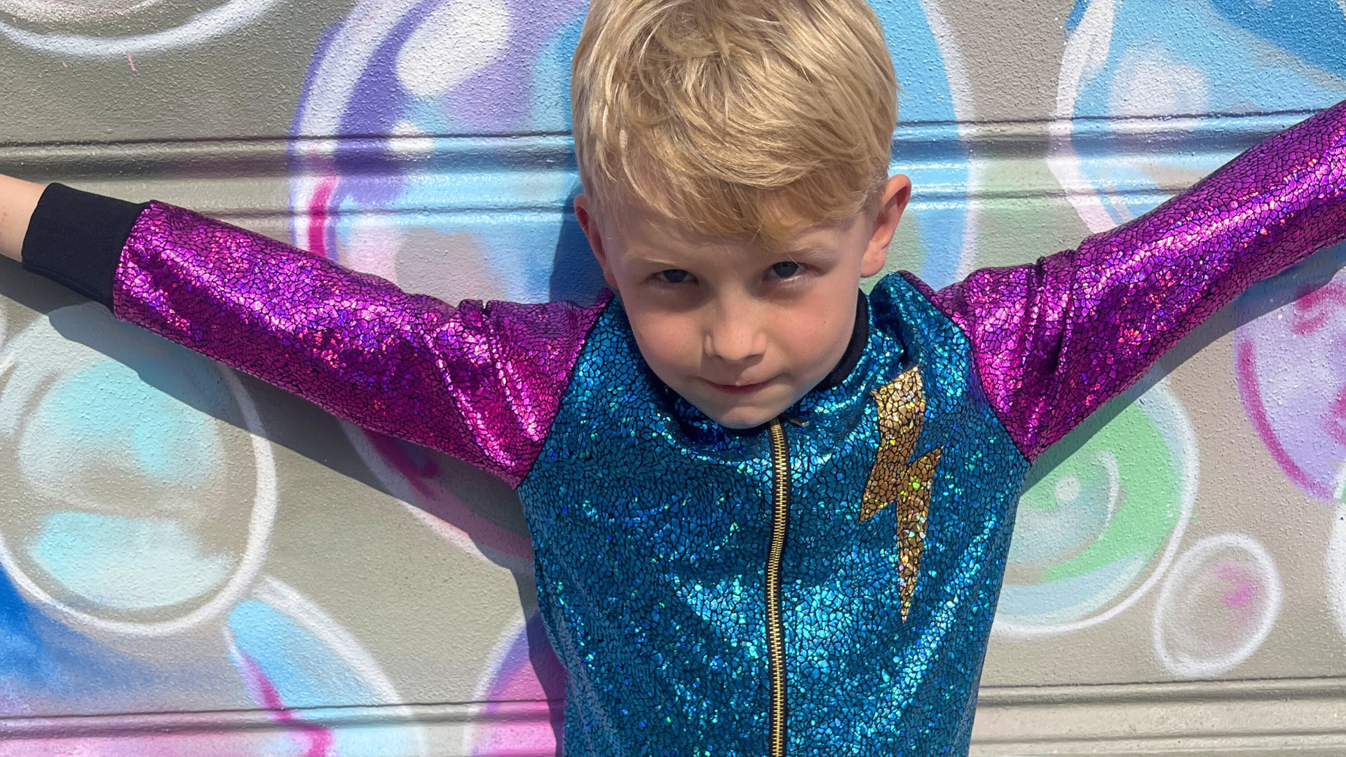 Child wearing a blue and purple sequin outfit with arms outstretched in front of a colorful bubble background.